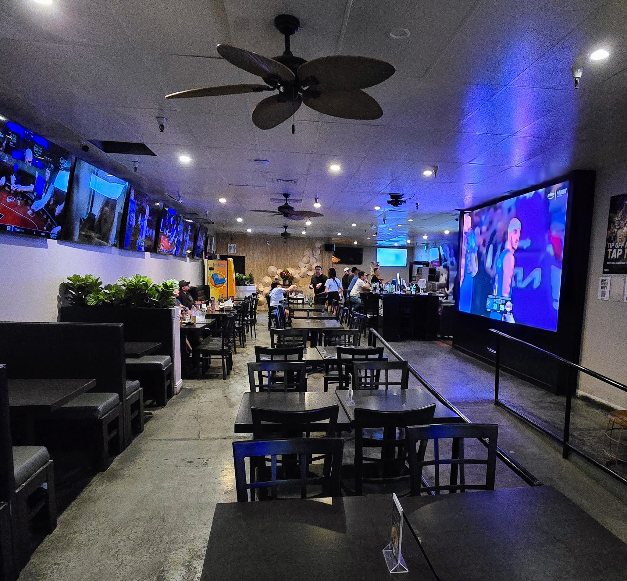 Inside Nhà Hotspot — TV wall lit up with the game, fiddle-leaf fig in the booth, full bar in the back